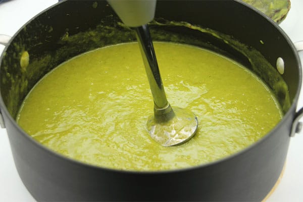 Creamy asparagus soup is pureed with an immersion blender in black soup pot.