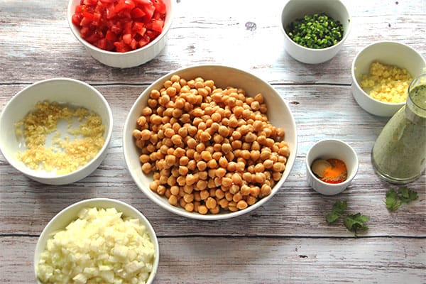 chickpeas, chopped onions, minced garlic and ginger, diced tomatoes, diced jalapenos, coriander cream and spices in white dishes on board.