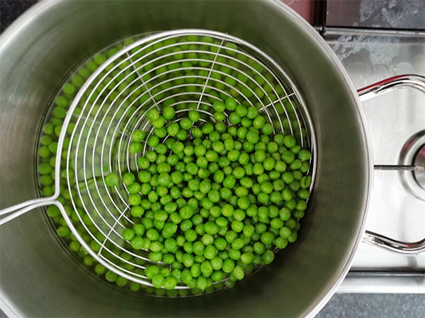 Frozen peas are simmered in pot and some are held up in a hand strainer.