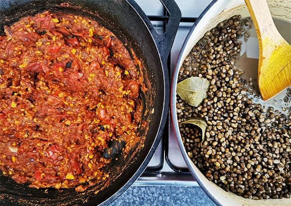 overhead view of black pan with tomato, onion, garlic, ginger and spices for tempering in black pan and alongside, urid beans (whole black lentils, with bay leaves in another pot all on stove top.