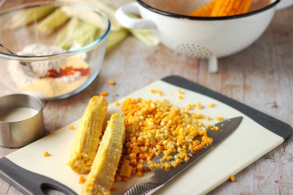 Fresh corn kernels are removed from cobs on white cutting board with bowl of dry ingredients and plant milk.