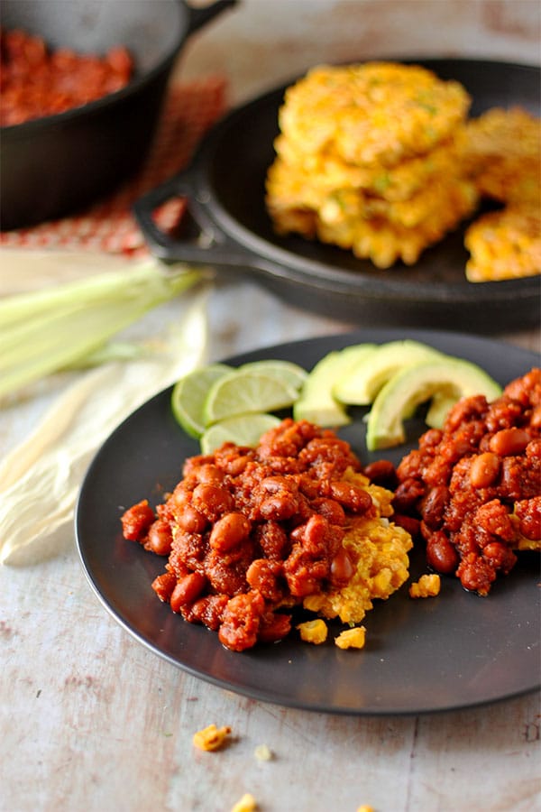 2 corn fritters smothered with red, chipotle baked beans with avocado and lime slices with fritters and more beans in background.