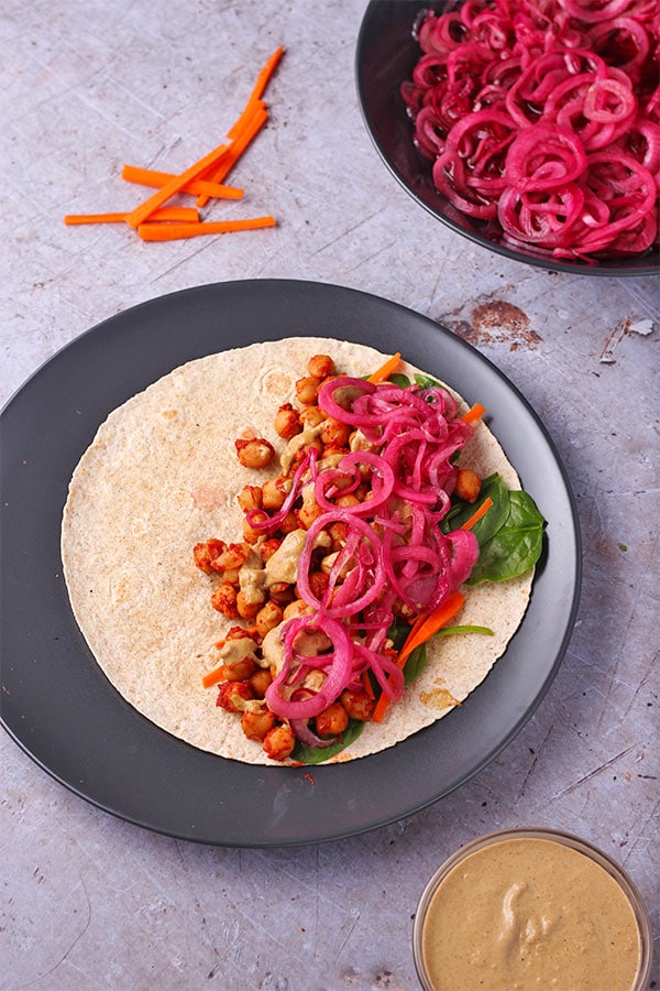 BBQ chickpea wraps, spinach, carrot sticks, marinated red onions and creamy tahini dressing on black plate with dressing and onions in separate bowls.