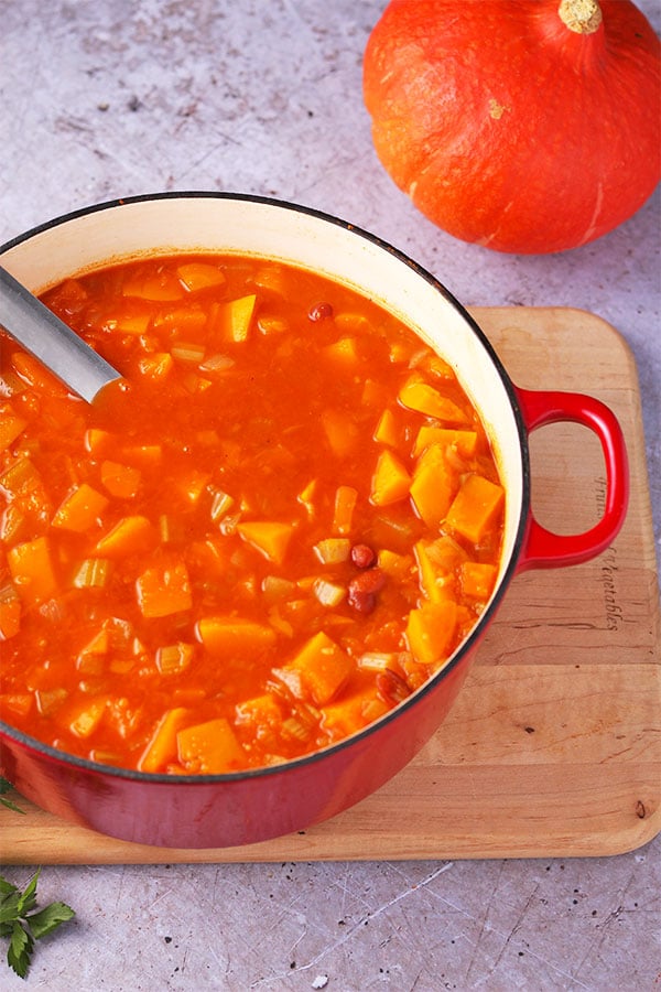 healthy pumpkin soup with diced, fresh pumpkin, carrots and vegetables in red pot on board.