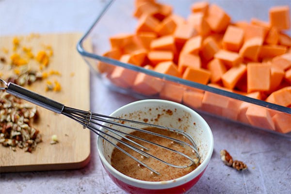 sauce for maple pecan sweet potatoes in red and white bowl with whisk, board with chopped pecans and orange zest and clear glass baking dish with diced sweet potatoes.