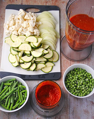 curry paste, frozen green beans and peas, blender with tomato puree, and chopping board with sliced onions, zucchini and cauliflower florets.
