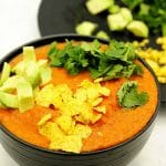 Easy vegan tortilla soup in black bowl with tortilla chips, avocado and coriander on top and plate behind with garnishes