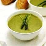 creamy asparagus soup in 2 white bowls with bread in the background.
