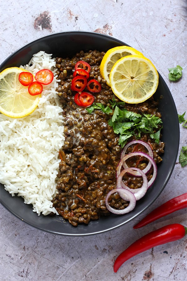 Dal Makhani - black lentil dal in white bowl with white rice garnished with sliced red chilis, lemon slices and cilantro with red chili peppers and chopped cilantro on stone board.