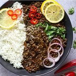 Dal Makhani - black lentil dahl in black bowl with white rice, red onion slices, lemon slices and chopped cilantro on stone board with whole red chilis. Recipe title in black text in white box on picture with Another Music in a Different Kitchen logo.