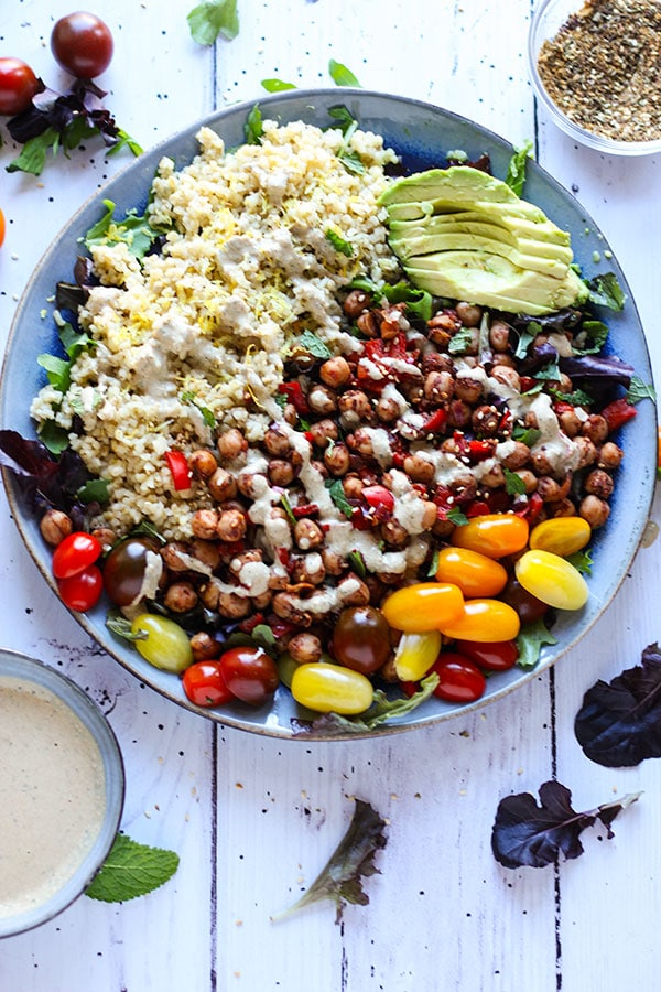 Middle Eastern chickpea salad on plate with salad greens, chickpeas, bulgur, cherry tomatoes, sliced avocado with dish of dressing and dish of spice blend.