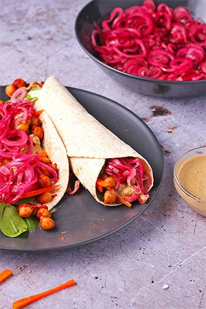 Black plate with BBQ chickpea wraps with marinated red onions, lettuce, carrots and creamy tahini dressing.