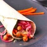 wrap with BBQ chickpeas, marinated red onions, creamy tahini dressing, carrot sticks and spinach on black plate.
