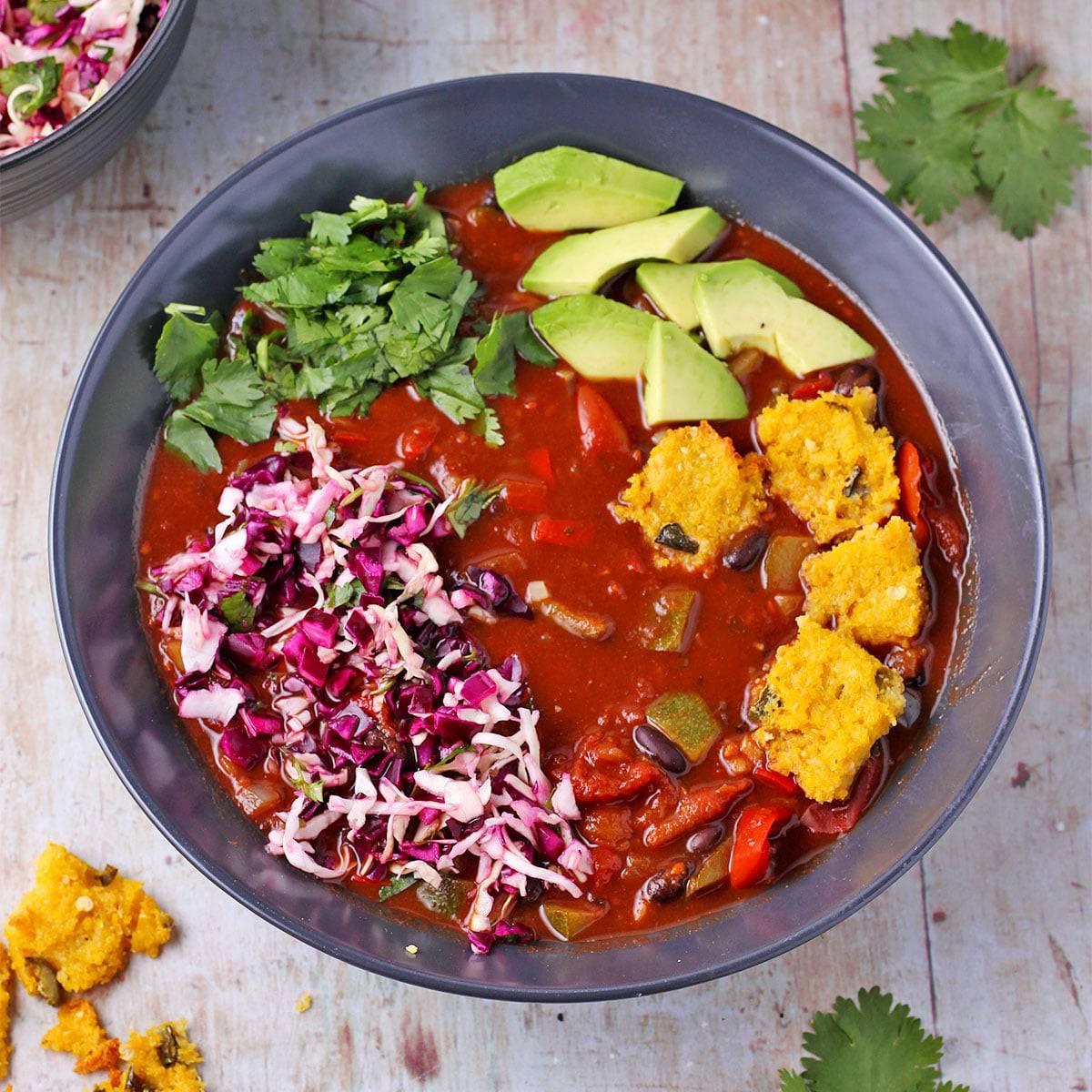 Overhead shot of bowl of black bean chili with cocoa powder topped with jalapeno polenta squares, cabbage slaw, chopped cilantro and avocado cubes