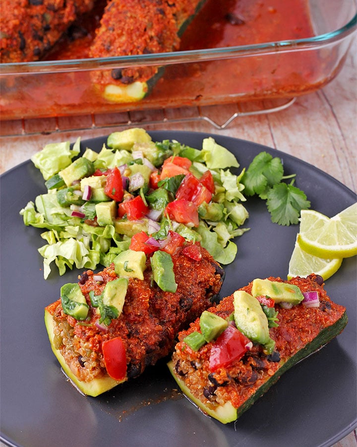 Mexican zucchini boats filled with quinoa and black beans and covered with enchilada sauce on black plate topped with avocado salsa with salad and salsa