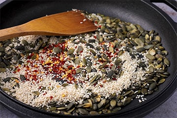 pepitas, sesame seeds and red chili flakes are toasted in skillet with wooden spoon