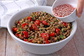 dressing with minced garlic, cranberry balsamic and lemon juice is poured over lentils, tomatoes and cilantro in white bowl.
