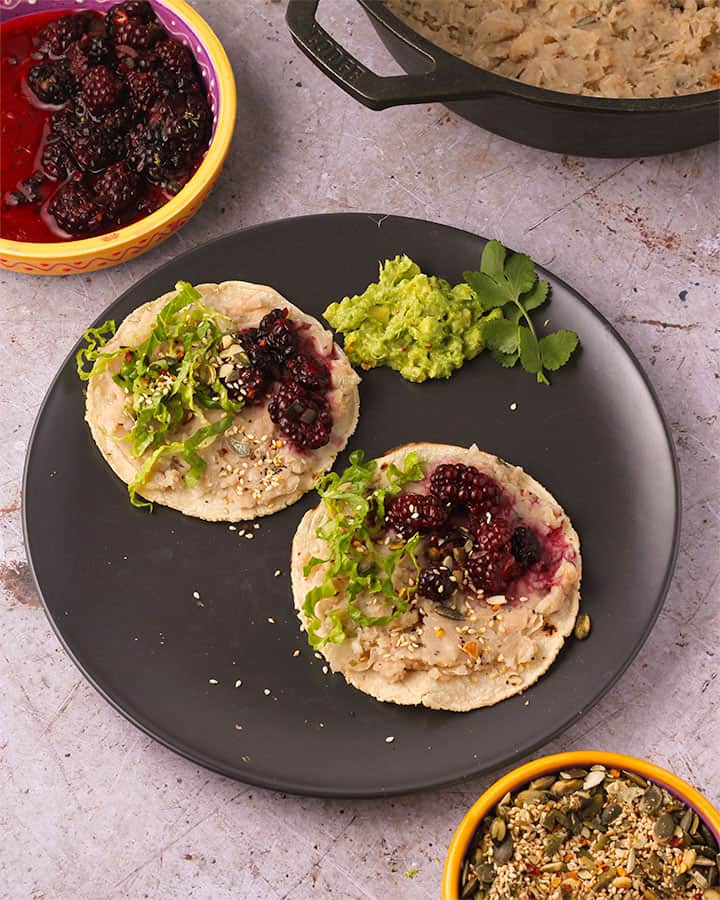 Overhead of toasted pepitas, sesame seeds and red chili in bowl with tostadas with refried white beans and blackberry salad and shredded lettuce on black plate with bowl of blackberry salsa and black pan of refried white beans