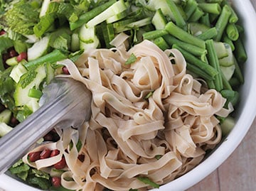 White rice noodles are added with tongs to bowl with green beans, bean sprouts, cucumbers, scallions and chopped mint, cilantro and basil.
