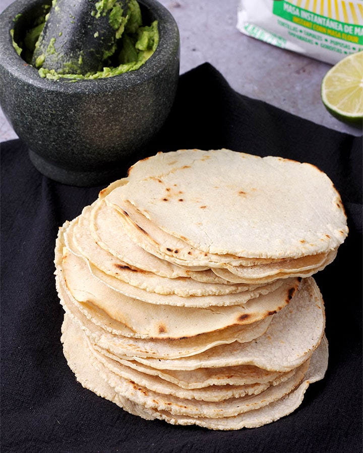 Corn tortillas made with masa harina are stacked on black cloth with mortar of smashed avocado in background.