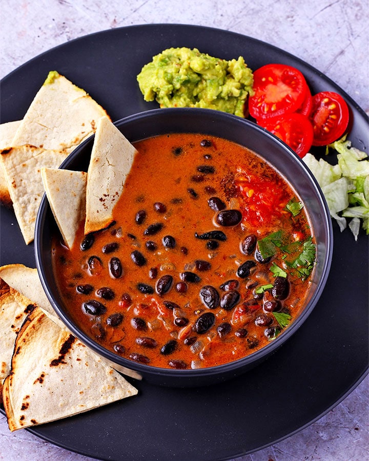overhead of saucy chipotle black beans in black bowl with 2 tortilla chips in bowl