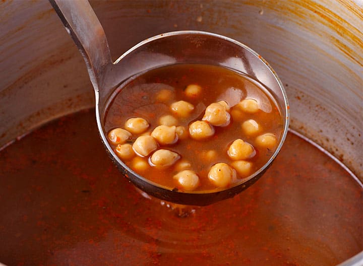 Ladle of cooked chickpeas and spices in broth is held over cooking pot.