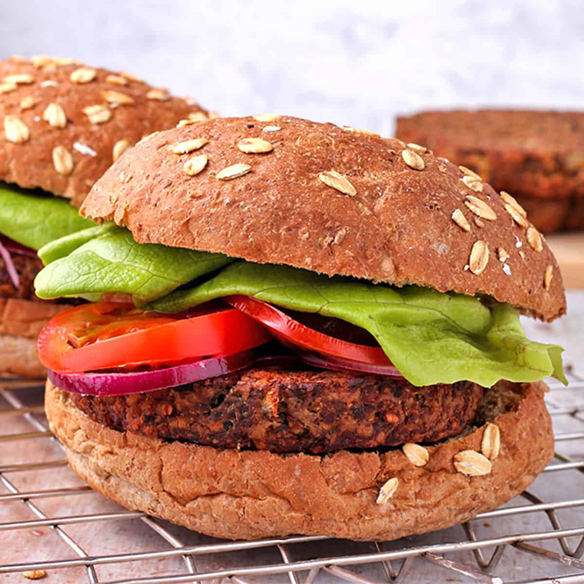 burgers made with tempeh are assembled with bun on bottom and top with red onion, tomato slices and lettuce and placed on a wire rack.