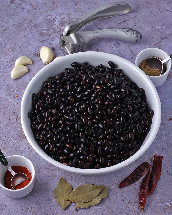 Dried black beans are placed in a white bowl with small white bowl of chili powder with tablespoon in it, 3 bay leaves, 3 dried red chilies, a small white bowl of ground cumin with teaspoon in it, 3 cloves of garlic and a gray garlic press