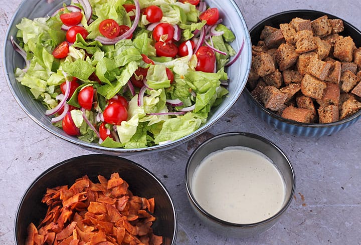 coconut bacon in black bowl, white Caesar dressing in blue bowl, croutons in blue bowl and lettuce, sliced tomatoes and sliced red onion in larger blue bowl