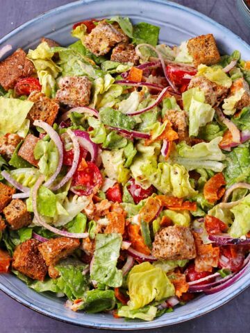 Overhead shot of vegan Caesar salad with chopped lettuce, sliced cherry tomatoes, sliced red onions, coconut bacon, croutons and dressing in blue bowl