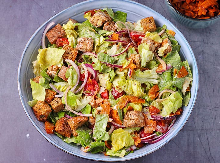overhead shot of vegan Caesar salad with coconut bacon, croutons, lettuce, cherry tomatoes, red onions and dressing in large blue bowl.
