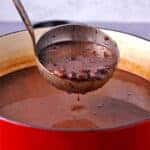 A red soup pot is filled with creamy black bean soup and a ladle filled with soup is lifted over the pot.