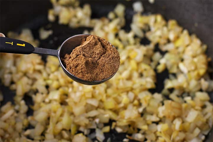 spoonful of garam masala held over sauteed onions, ginger and garlic in black pan