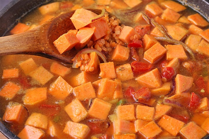 Diced sweet potatoes are cooked in pot with tomatoes, broth and red lentils and stirred with wooden spoon.