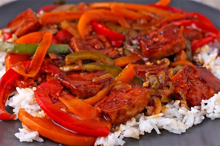 sliced peppers, onions, baked tofu cubes with red sweet and sour sauce over white rice on black plate