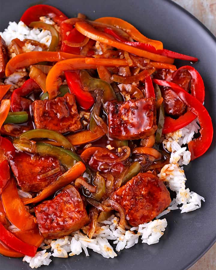 sweet and sour tofu with pepper and onion slices over white rice on black plate