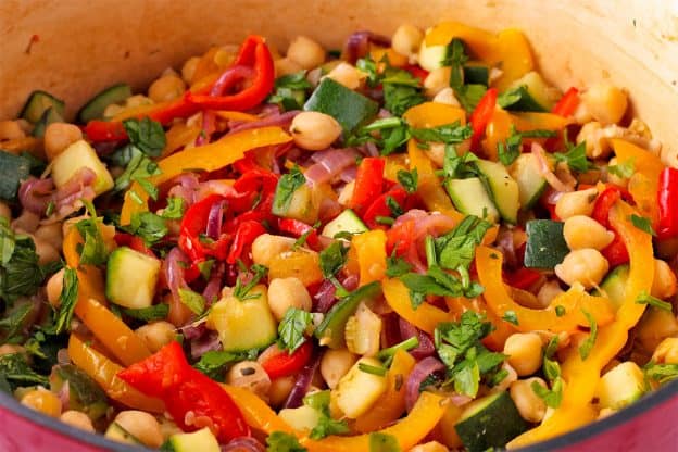 Sliced yellow and red peppers, sliced red onions, diced zucchini and chickpeas with parsley in red pot.