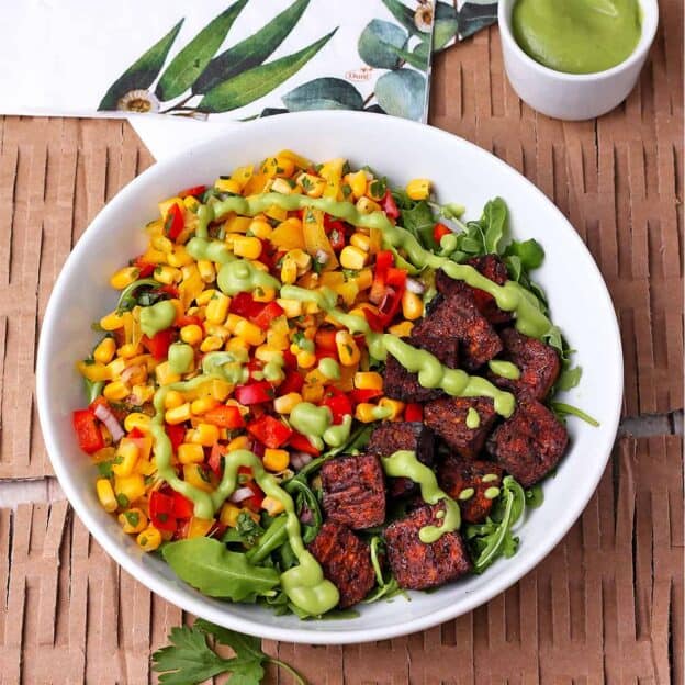 baked tofu pieces in a white bowl with arugula, corn relish and avocado dressing. 