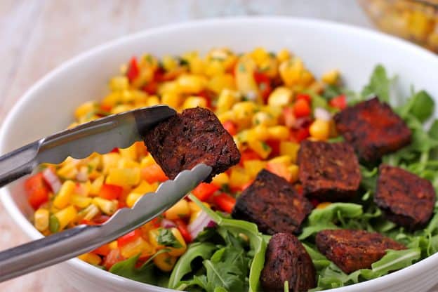 A baked tofu piece is placed on a bowl of arugula and corn relish