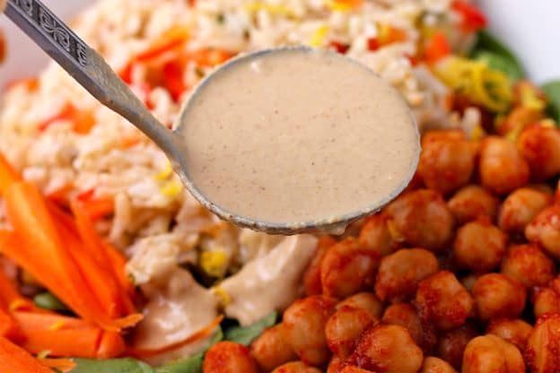 ginger-tahini dressing in a ladle is added to buffalo chickpeas, carrot sticks, spinach, and rice.