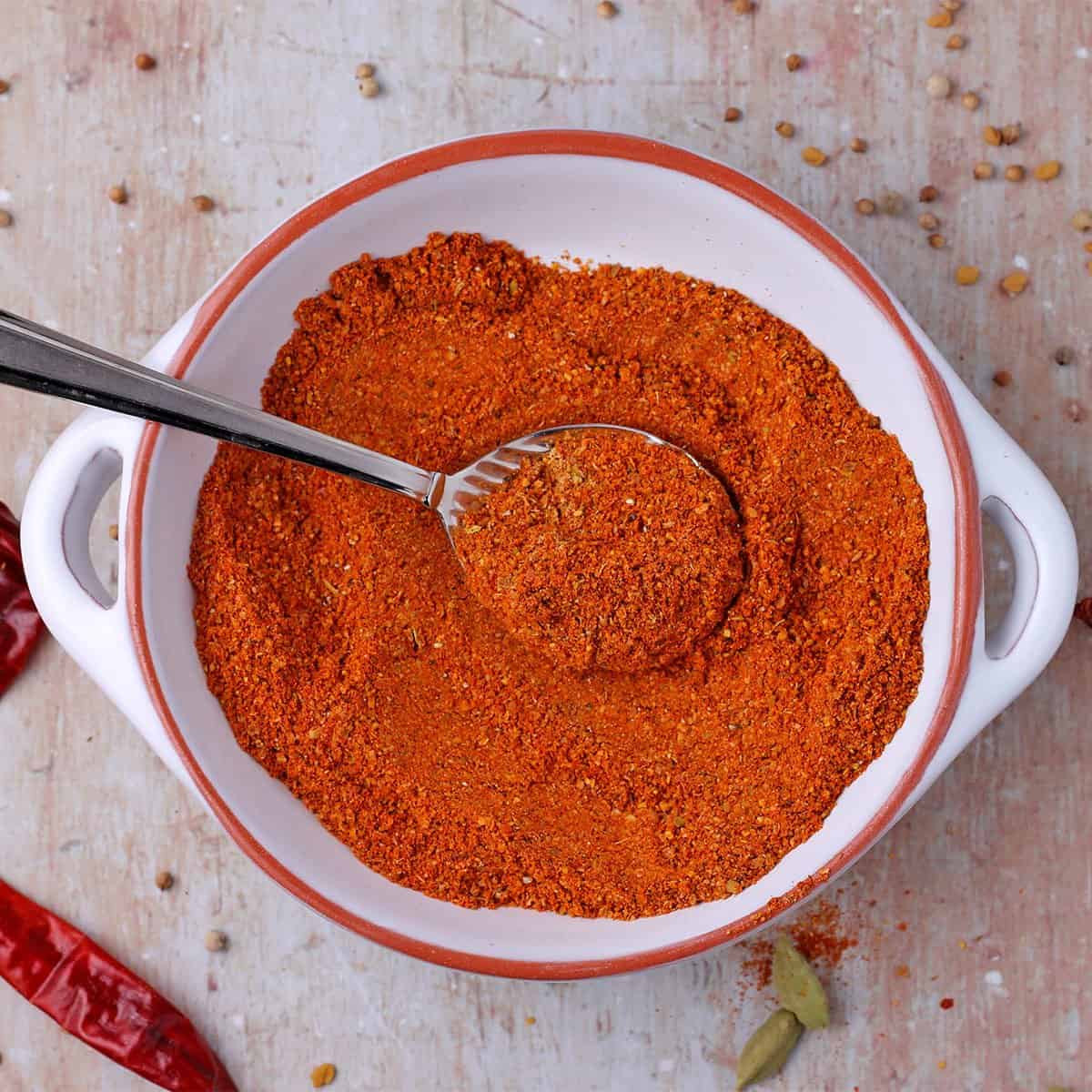 bowl of red berbere spice blend with dried red chilies, cardamom pods, fenugreek seeds, and coriander seeds.