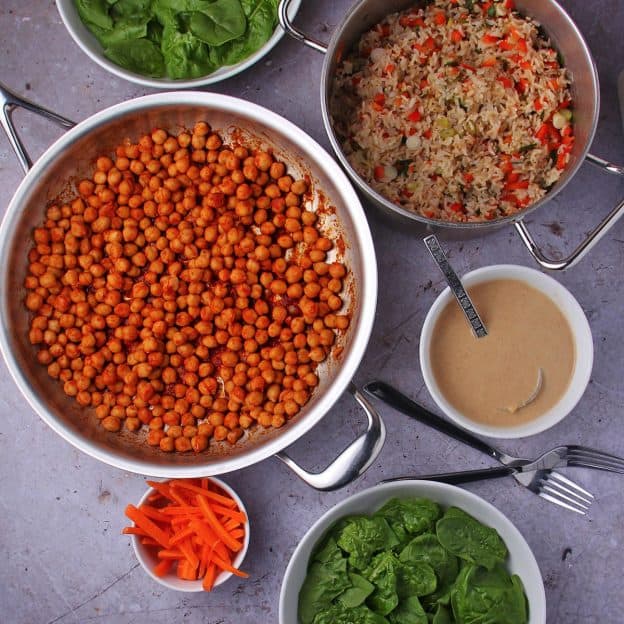 bowl of fresh spinach, bowl of carrot sticks, pan of buffalo chickpeas, bowl of ginger-tahini dressing, and pan of brown rice with red peppers