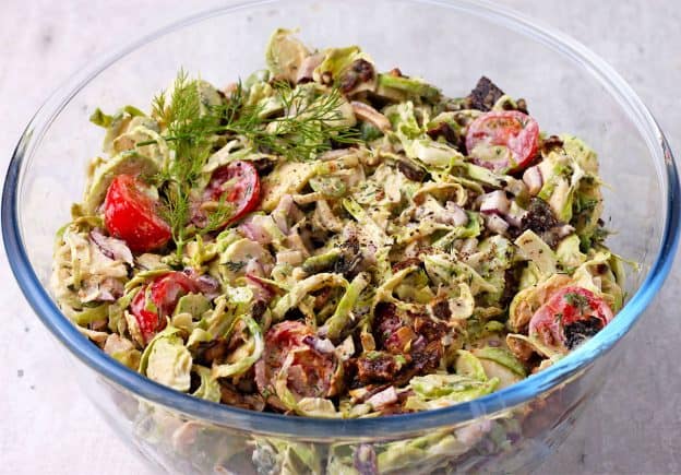 glass bowl with shaved Brussels sprouts, sliced tomatoes, red onion, vegan bacon, and cashew ranch dressing