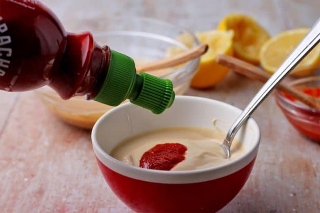 Sriracha is added from a bottle to a red bowl with tahini.