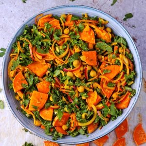 A blue salad bowl is filled with roasted sweet potatoes, chopped kale, turmeric chickpeas, and red onions.