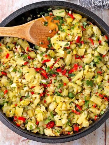 A black cast iron skillet is filled with pineapple chutney with cumin seeds, red chilies, cilantro, onion, ginger, and diced pineapple.