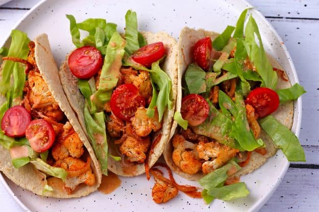 3 roasted cauliflower tacos with Chipotle cream, lettuce and tomatoes on a white plate.