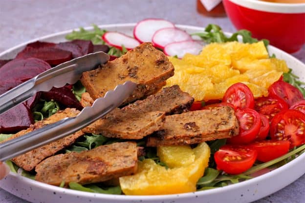 A baked tofu slice is added with tongs to a plate of orange segments, cooked beetroot, cherry tomatoes, radishes, and arugula.
