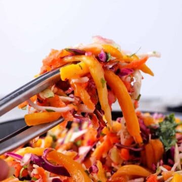 Tongs hold coleslaw with green and red cabbage, carrots, chopped cilantro, and mango strips over a bowl of slaw.