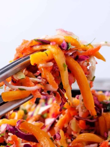 Tongs hold coleslaw with green and red cabbage, carrots, chopped cilantro, and mango strips over a bowl of slaw.